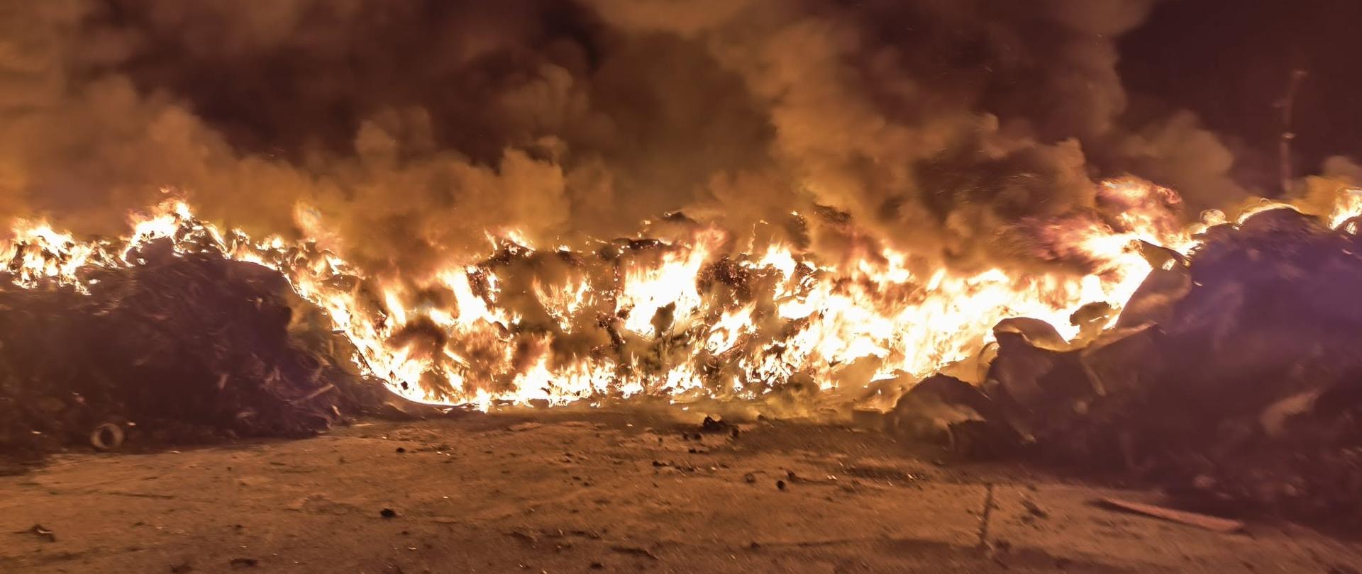 Noc, dużo dymu, na pierwszym planie dużo ognia, pali się hałda czarnego materiału na placu