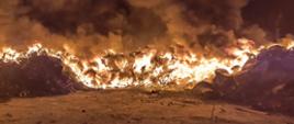 Noc, dużo dymu, na pierwszym planie dużo ognia, pali się hałda czarnego materiału na placu