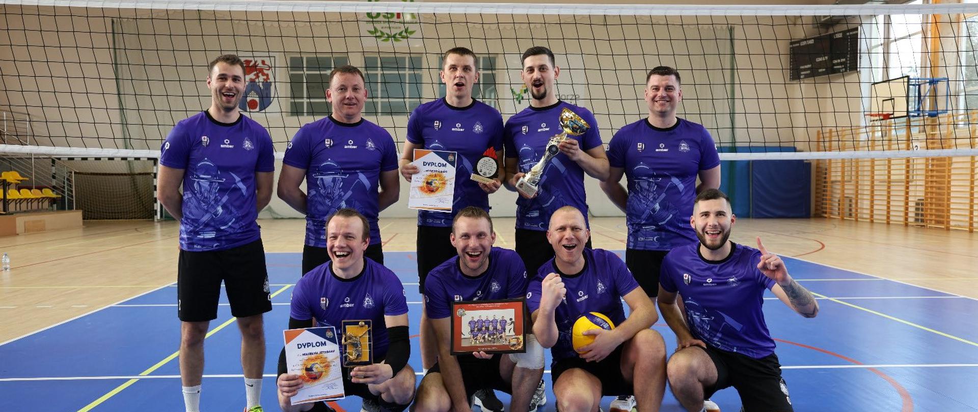 Grupa mężczyzn w sportowych koszulkach stoi w hali sportowej przy siatce do siatkówki. Zawodnicy pozują do zdjęcia po zdobyciu nagród – trzymają puchar, dyplomy i statuetki. Część z nich stoi, a część klęczy na parkiecie.