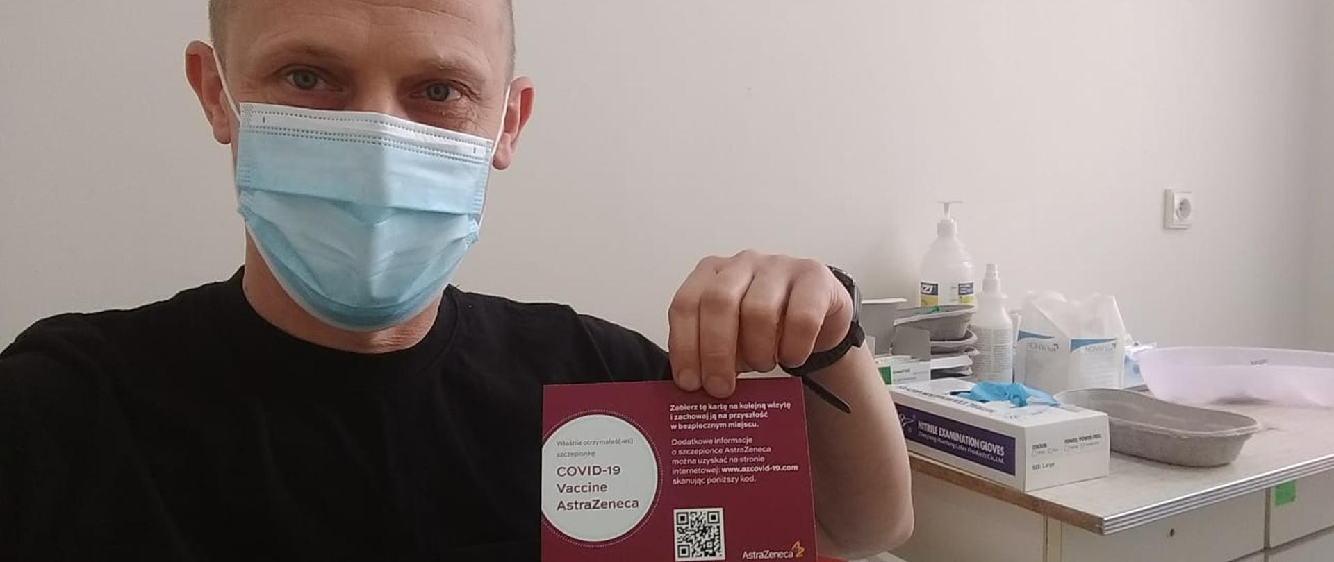 Zdjęcie przedstawia strażaka znajdującego się w gabinecie szczepień, który w lewej dłoni trzyma kartę szczepienia przeciwko COVID-19.