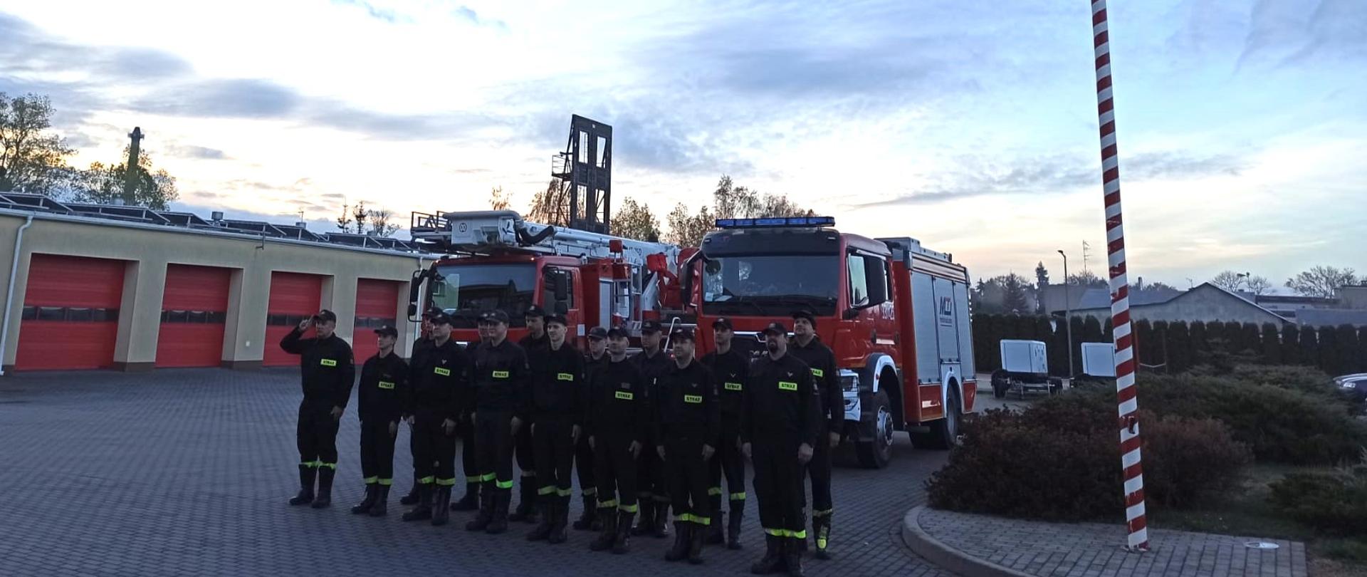 Zdjęcie przedstawia strażaków z JRG w Pruszczu Gdańskim stojących w dwuszeregu podczas uroczystej zmiany służby na tle dwóch samochodów strażackich, obok masztu flagowego z podniesioną flagą państwową.