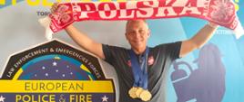 Mężczyzna z trzema medalami na szyi nad głową trzyma w dłoniach szalik z napisem Polska, w tle baner zawodów European Police & Fire Games