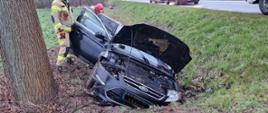 Widać samochód osobowy w rowie, w pobliżu strażacy, po lewej drzewo