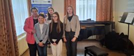 Sala kameralna Uczniowie stoją wspólnie z nauczycielem pozując do zdjęcia, w tle czarny fortepian, baner reklamy szkoły, z boku i z tyło dwa duże okna z brązowymi zasłonami. Po prawej stronie krzesło do fortepianu oraz pulpit z nutami.