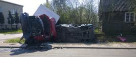 Na zdjęciu widać uszkodzone pojazdy, osobowy i dostawczy. Po prawej stronie widać podłogę samochodu osobowego, po lewej zniszczony przód samochodu dostawczego. Po lewej stronie zdjęcia widoczny jest budynek mieszkalny, piętrowy, w kolorze szarym. Po prawej opuszczony dom drewniany z metalowym płotem przed. Na pierwszym planie widać asfalt pasa jezdni. Za pojazdami zielone krzewy.