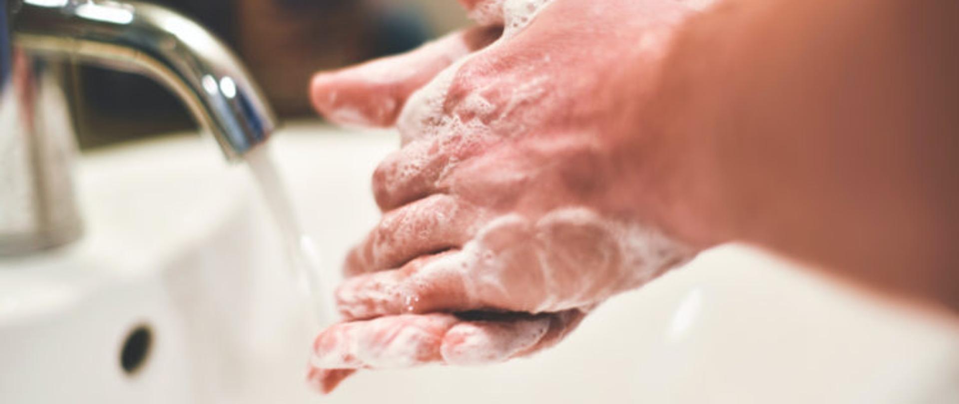 zdjęcie przestawiające mycie rąk, na głównym planie biała porcelanowa umywalka z srebrnym kranem, woda leci nieprzerwanym strumieniem. nad zlewem widoczne dłonie całe w pianie, jedna dłoń na drugiej, 