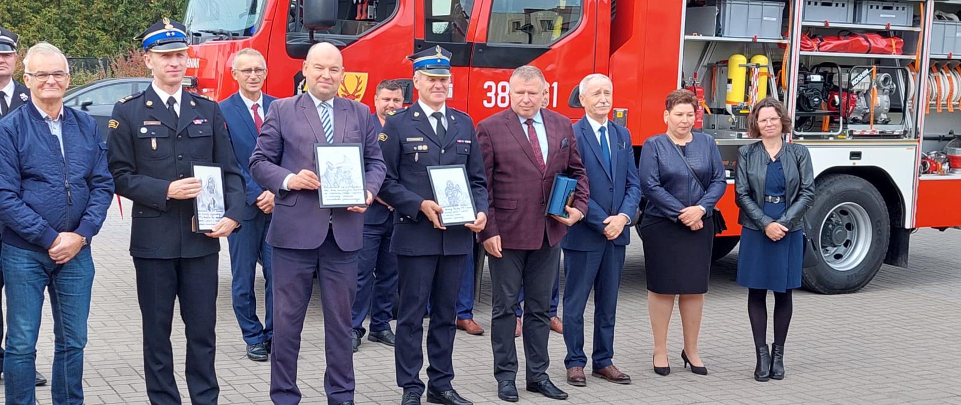 grupa mężczyzn i kobiet stoi przed samochodem strażackim, kilku z nich to strażacy, trzech mężczyzn trzyma w ręku dyplomy