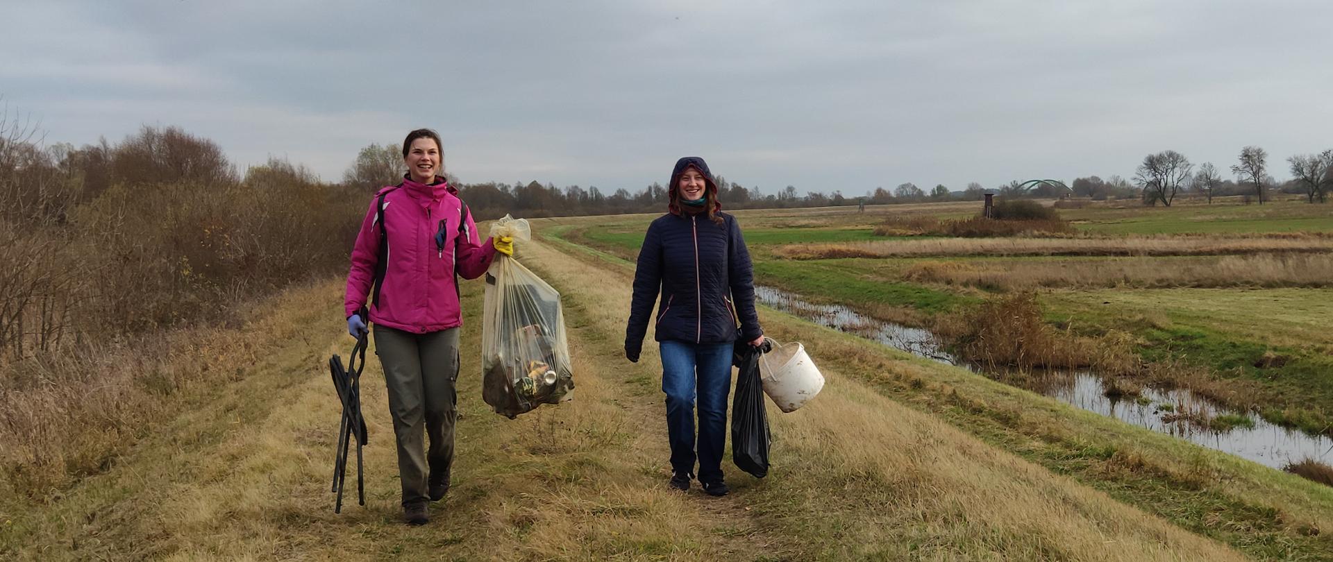 Dwie młode kobiety w kurtkach idą i niosą worki z odpadami. Uśmiechają się. Idą polną drogą. Po prawej stronie widoczny niewielki ciek wodny. Na niebie szare chmury. 