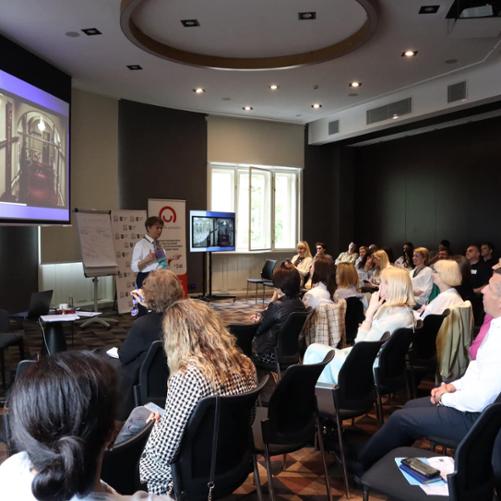 Sala konferencyjna, na krzesłach siedzą uczestnicy spotkania, przed nimi stoi kobieta prowadząca szkolenie, a za nią baner z logo Polska pomoc