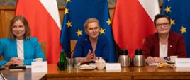 Duża sala ze ścianami w zielony wzorek, przy drewnianym stole siedzi od lewej: Szefowa Gabinetu Politycznego Ada Guźniczak, minister Barbara Nowacki i wiceminister Katarzyna Lubnauer. Na stole rozłożone notatki oraz ustawione napoje. W tle flagi Polski i UE