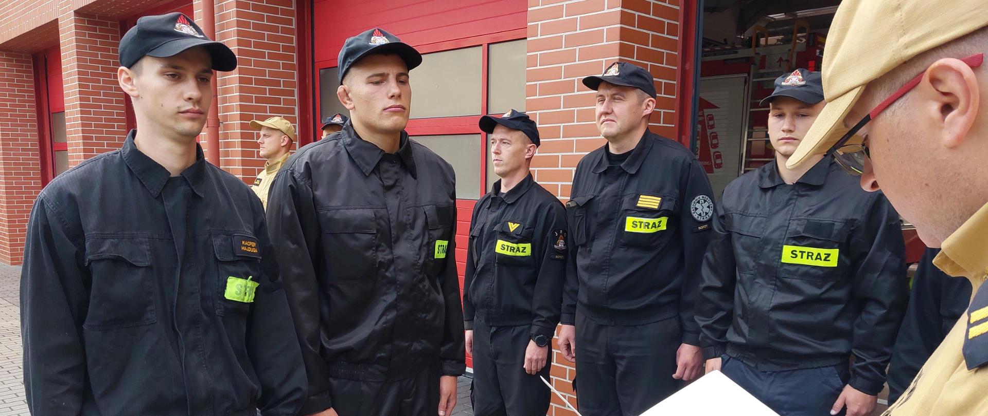 Dwóch strażaków stoi przed zmianową służbową w trakcie odczytywania aktów mianowania przez komendanta miejskiego.