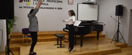Na zdjęciu widnieje nauczyciel fortepianu mgr Dorota Skibicka, która w auli szkoły, obok czarnego fortepianu wykonuje wraz z uczniem ćwiczenia gimnastyczne. Tło zdjęcia jest białe i jasno brązowe.