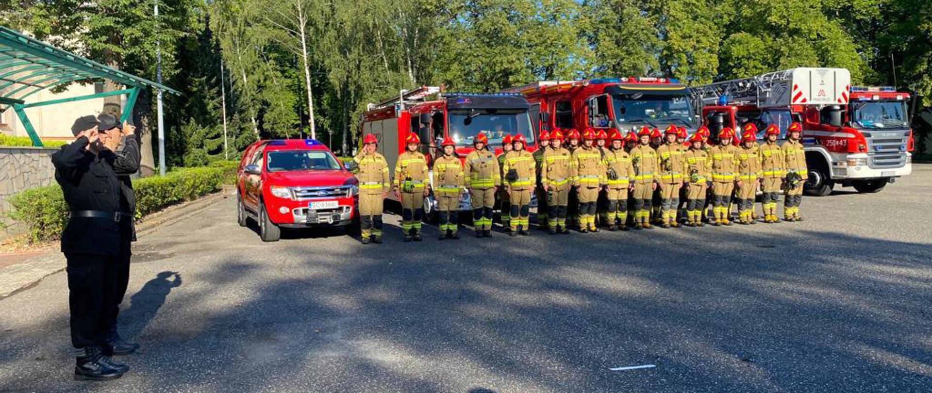 Plac wewnętrzny Centralnej Szkoły PSP, na którym stoją na baczność strażacy podczas minuty ciszy, za nimi samochody z włączoną sygnalizacją świetlno - dźwiękową