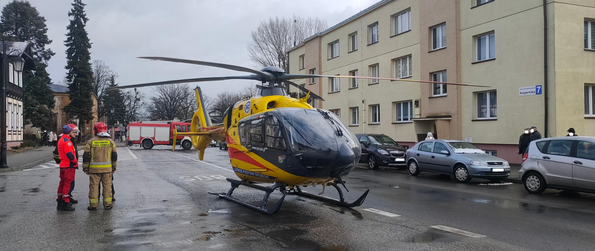 Na środku zdjęcia widać helikopter lotniczego pogotowia ratunkowego koloru żółto-czarno-czerwonego, stojący na ulicy asfaltowej. Droga jest mokra, padał deszcz. W tle za helikopterem widać czerwony samochód strażacki blokujący przejazd ulicą. Po lewej stronie zdjęcia stoją pilot śmigłowca oraz strażacy JRG Ciechocinek i policjant. Wzdłuż drogi zaparkowane są trzy samochody osobowe koloru czarnego, ciemno szarego i szarego. Po prawej stronie przy ulicy widać trzykondygnacyjny budynek mieszkalny wielorodzinny koloru piaskowego z brązowymi rynnami. Na chodniku przed budynkiem stoją ludzie przyglądający się akcji. Po lewej stronie widać fragment innych zabudowań oraz drzewa. Pogoda jest pochmurna.