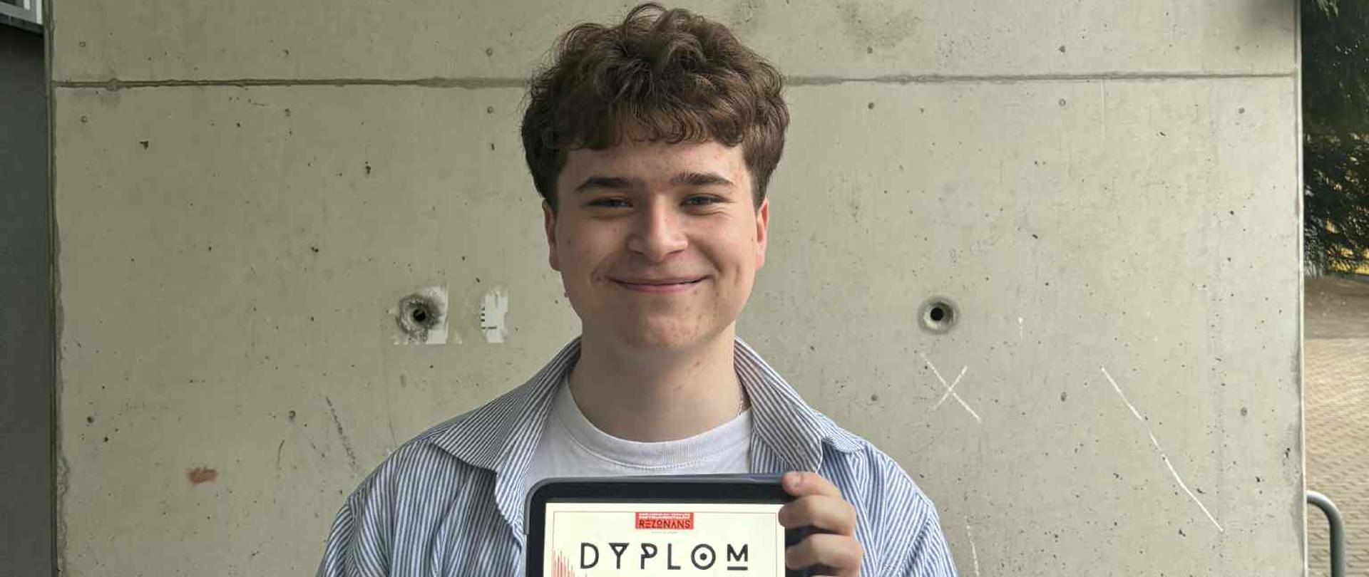 Uczeń ZPSM z dyplomem w ręku, na tle ściany i godła Polski oraz nazwy szkoły