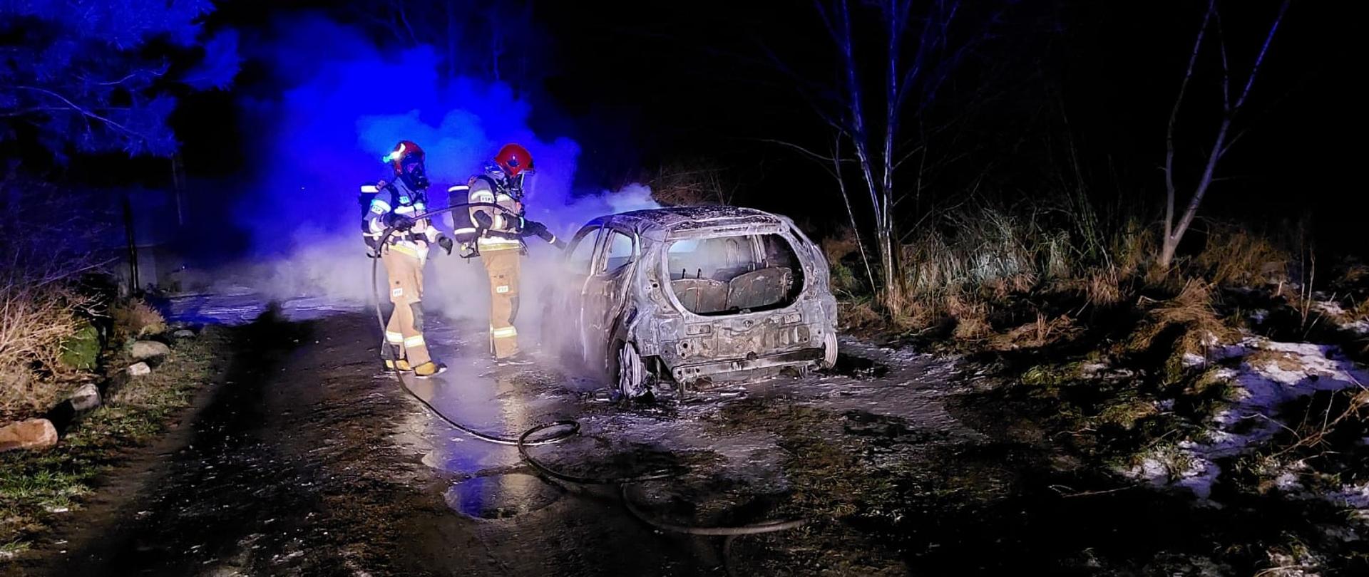 Zdjęcie przedstawia spalony samochód osobowy zaparkowany na drodze wewnętrznej. Dwóch strażaków zabezpieczonych w aparaty ochrony układu oddechowego gasi pożar samochodu. Samochód całkowicie spalony. Pora nocna. 