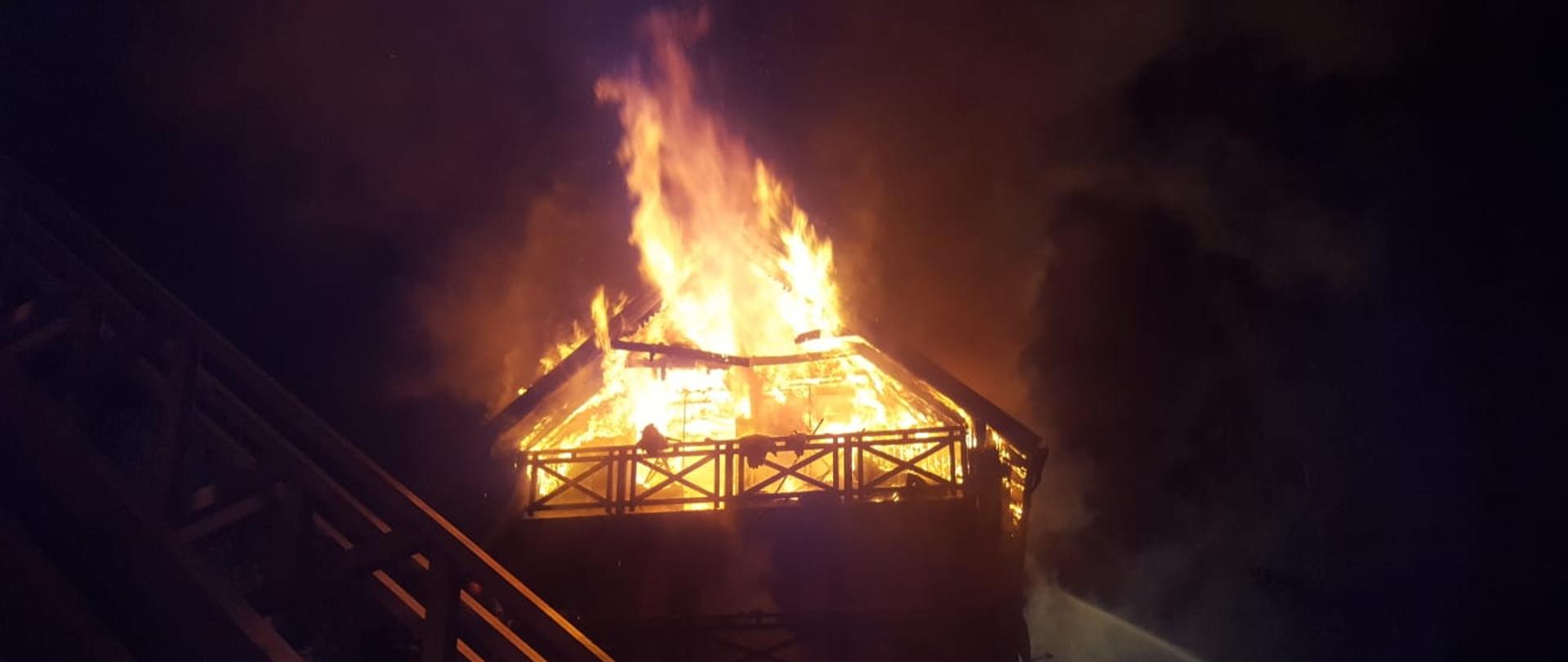 Zdjęcie przedstawia dom letniskowy objęty pożarem, ogień wychodzi przez wszystkie otwory na poddaszu.