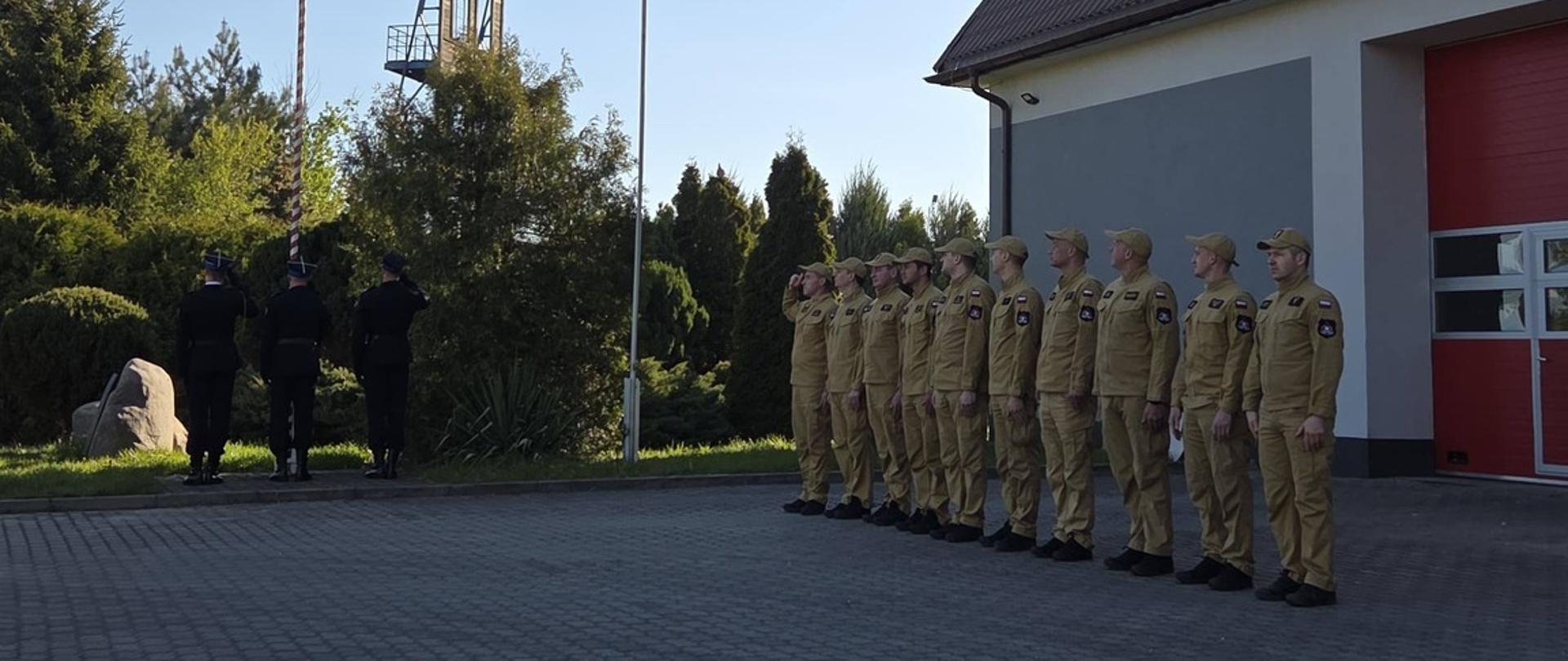 Na zdjęciu widać grupę strażaków stojących w szeregu przed budynkiem z czerwonymi bramami garażowymi. Obok masztu z polską flagą odbywa się uroczystość podniesienia flagi, podczas której kilka osób stoi na baczność i salutuje.