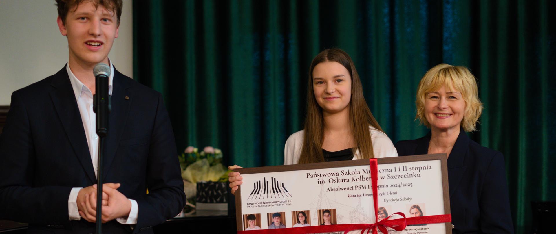 Na zdjęciu troje osób podczas uroczystości szkolnej – młody mężczyzna w garniturze przemawia do mikrofonu, obok niego dziewczyna i kobieta w eleganckim stroju trzymają pamiątkową tablicę z fotografiami absolwentów Państwowej Szkoły Muzycznej I i II stopnia im. Oskara Kolberga w Szczecinku. W tle widoczne są zielone zasłony i kwiaty na fortepianie.