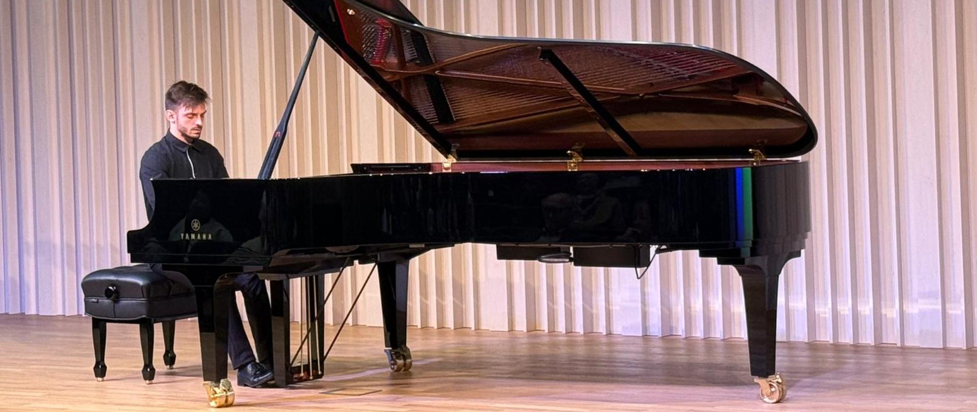 Pianista siedzi przy fortepianie koncertowym na scenie jasnej sali koncertowej. Gra w skupieniu, a przed nim siedzi publiczność zajmująca wszystkie rzędy krzeseł. W tle widoczne są pionowe, jasne panele akustyczne.