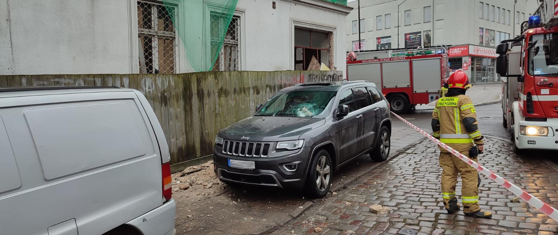 Budynek ogrodzony płotem przed budynkiem zaparkowane dwa samochody. W jednym z samochodów widać uszkodzenie przedniej szyby. prawa część jest wygrodzona taśmą, za którą stoi strażak oraz samochody pożarnicze.