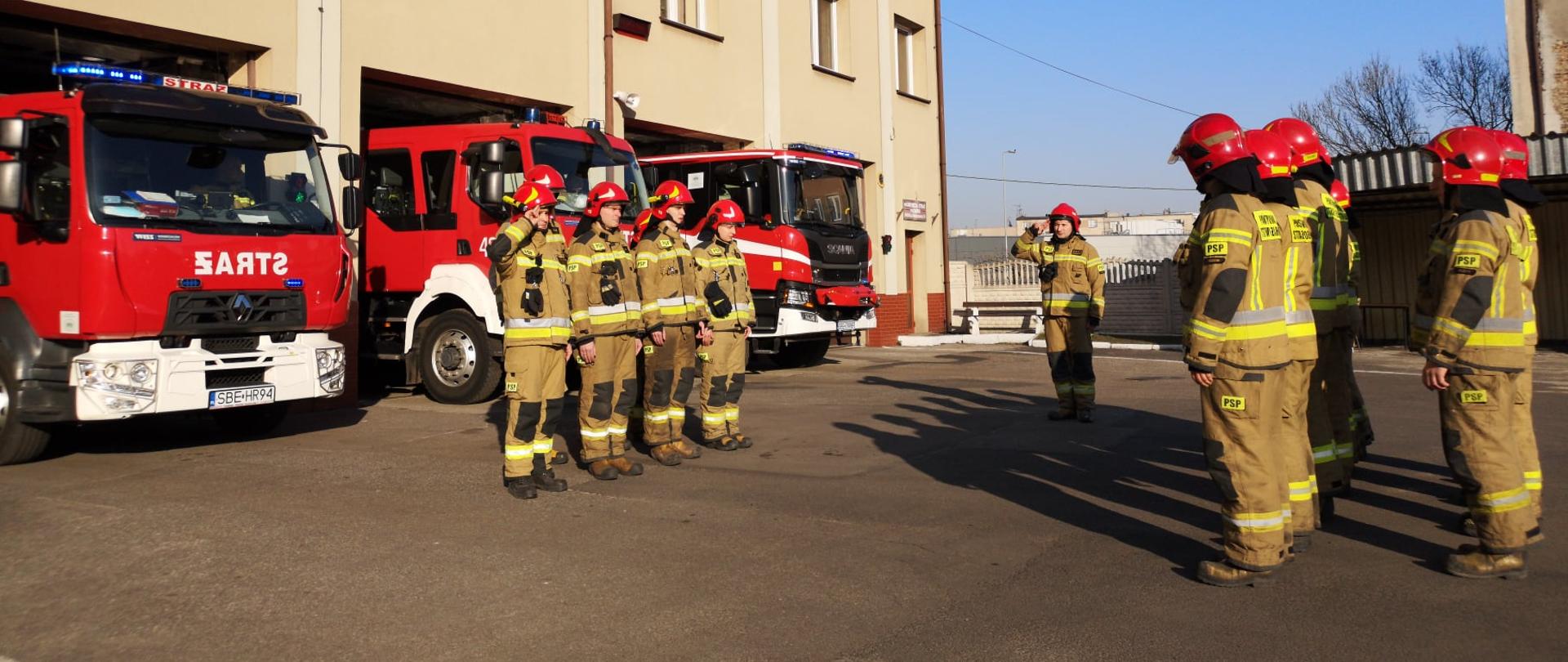 Zdjęcie ukazuję strażaków podczas zmiany służbowej na placu komendy przed garażami i samochodami ratowniczo gaśniczymi 