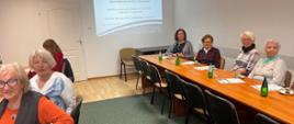 Kobiety siedzą przy stole na Sali konferencyjnej. Na stole stoją szklanki, woda w butelkach oraz ciasto. W tle na ścianie prezentacja