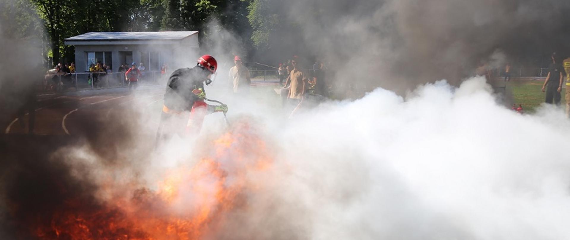 Zdjęcie obrazuje strażaka gaszącego pożar wanny, w której pali się łatwopalna ciecz. Jest to element rywalizacji strażaków podczas Mistrzostw Polski w Sporcie Pożarniczym.