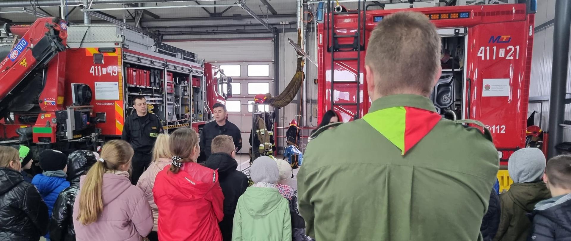 Strażacy na garażu JRG prezentują dzieciom sprzęt i pojazdy pożarnicze. Plecami do fotografa ustawiony jest harcerz w mundurze.