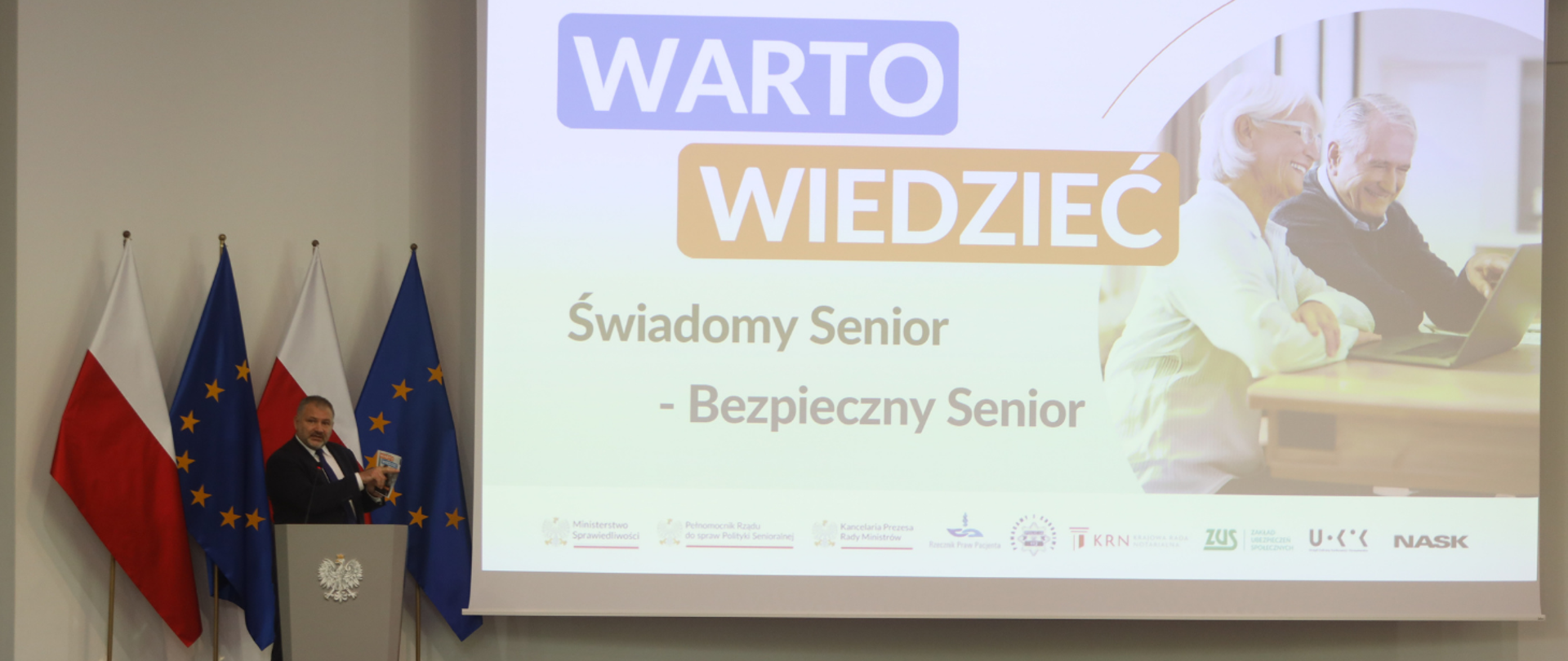 Minister w garniturze przemawia przy mównicy ozdobionej godłem Polski, stojąc na tle flag narodowych i unijnych. Za nim znajduje się duży ekran z napisem: „WARTO WIEDZIEĆ. Świadomy Senior - Bezpieczny Senior” oraz zdjęciem starszego małżeństwa przy komputerze. Prelegent trzyma w ręku małą książeczkę. 
