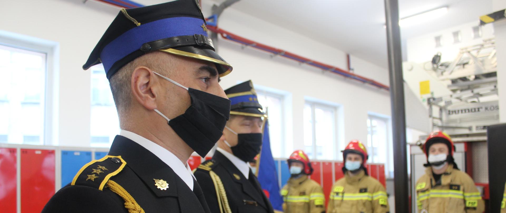 Zdjęcie przedstawia sylwetkę funkcjonariusza Państwowej Straży Pożarnej ubranego w mundur galowy w ujęciu z jego prawego profilu podczas uroczystego apelu z okazji przejścia na zaopatrzenie emerytalne. Na dalszym planie po lewej stronie od funkcjonariusza odchodzącego na zaopatrzenie emerytalne stoi komendant powiatowy państwowej straży pożarnej w lesku starszy brygadier Jacek Dydek. W tle stoi trzech strażaków systemu zmianowego ubranych w ubrania specjalne stojący w szeregu. Zdjęcie zrobione w garażu jednostki ratowniczo gaśniczej państwowej straży pożarnej w lesku.