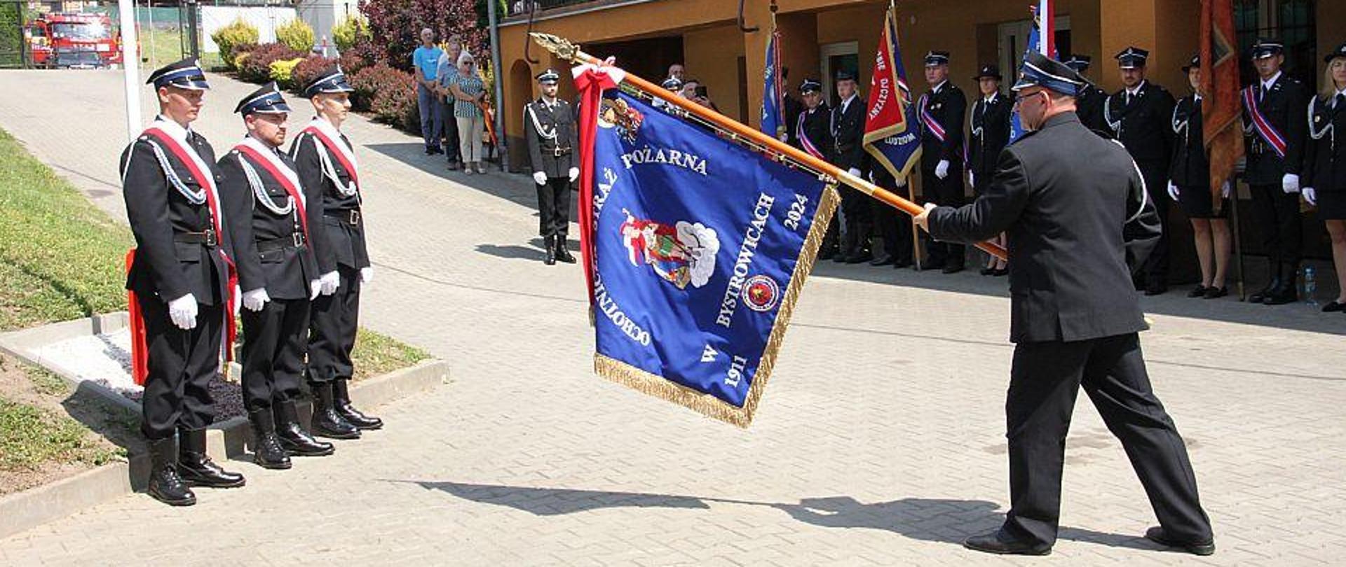 Zdjęcie przedstawia moment prezentowania sztandaru. W tle widoczny budynek oraz pozostałe poczty sztandarowe.