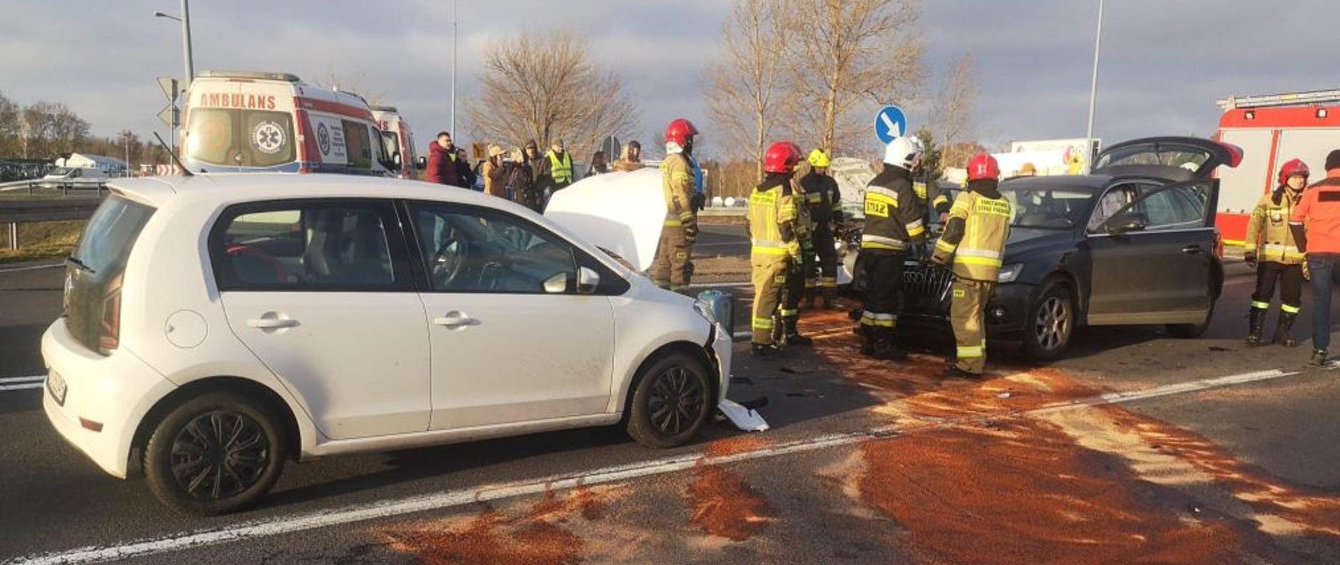Widok na miejsce zdarzenia. Centralny punkt zdjęcia stanowi białe rozbite auto. Na miejscu widać strażaków i wozy ratownicze.