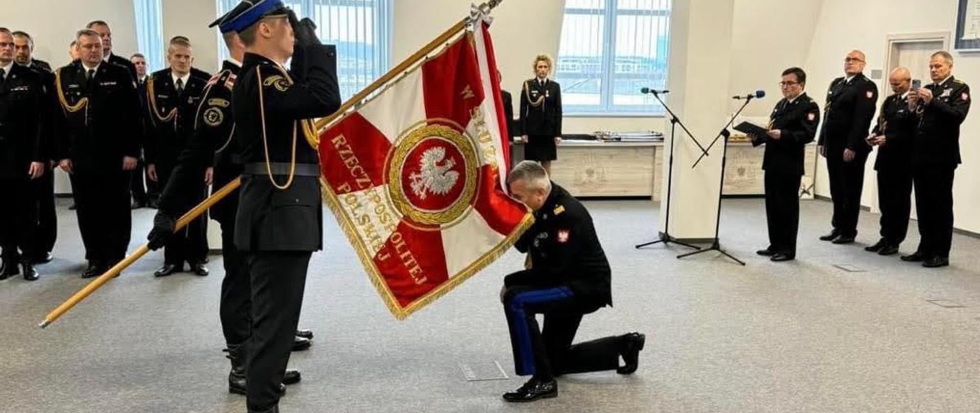 Funkcjonariusz Państwowej Straży Pożarnej klęczy przed sztandarem całując go sztandar trzyma poczet sztandarowy obok stoją strażacy w mundurach wyjściowych ze sznurem.