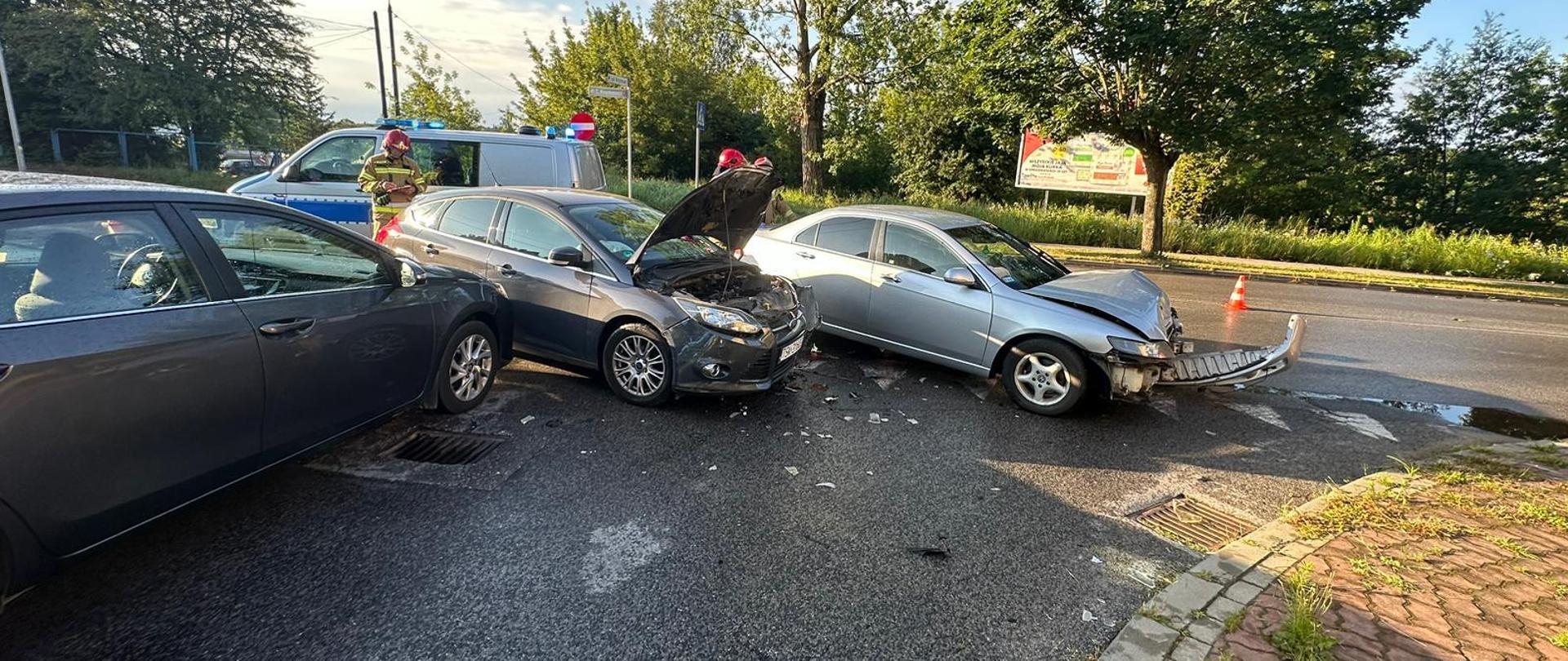 Trzy samochody osobowe po kolizji drogowej. Obok nich pracują strażacy i policjanci.