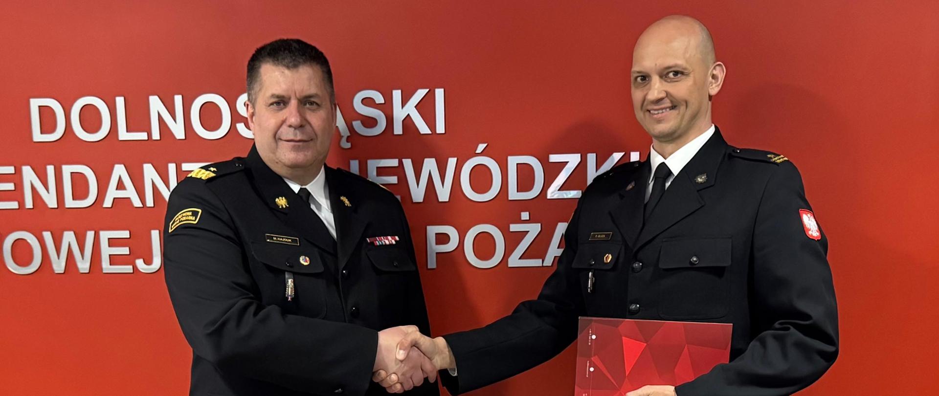 Zbliżenie na dwóch funkcjonariuszy Państwowej Straży Pożarnej w galowych mundurach, którzy uściskują sobie dłonie w oficjalnym geście na tle czerwonej ściany.