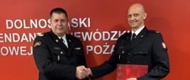 Zbliżenie na dwóch funkcjonariuszy Państwowej Straży Pożarnej w galowych mundurach, którzy uściskują sobie dłonie w oficjalnym geście na tle czerwonej ściany.