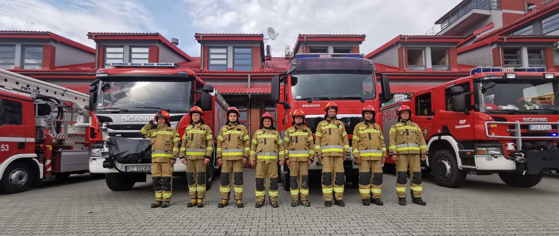 Zdjęcie wykonane na zewnątrz, przed drzwiami garażowymi jednostki ratowniczo – gaśniczej KP PSP w Sanoku. Na pierwszym planie stoi 8 strażaków w umundurowaniu bojowym. W tle 4 wozy bojowe
i budynek jednostki ratowniczo – gaśniczej.
