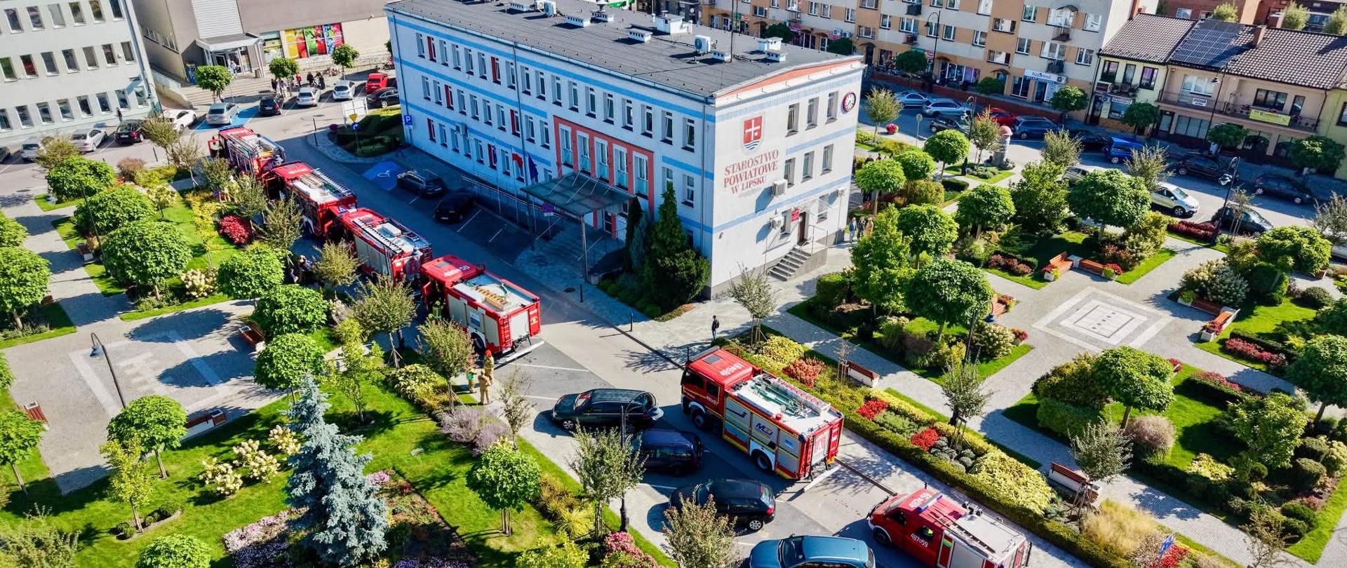 Widok z lotu ptaka na budynek Starostwa Powiatowego w Lipsku oraz przyległy plac i park. Przed budynkiem zaparkowanych jest kilka czerwonych samochodów strażackich w kolumnie, a wokół widoczne są tereny zielone, alejki spacerowe i budynki miejskie.