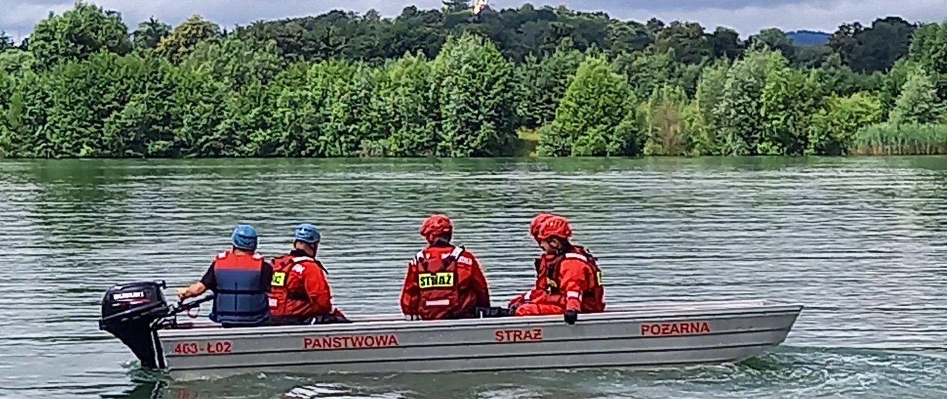 Strażacy w łodzi ćwiczący na zbiorniku wodnym ratownictwo wodne. W oddali brzeg porośnięty drzewami.