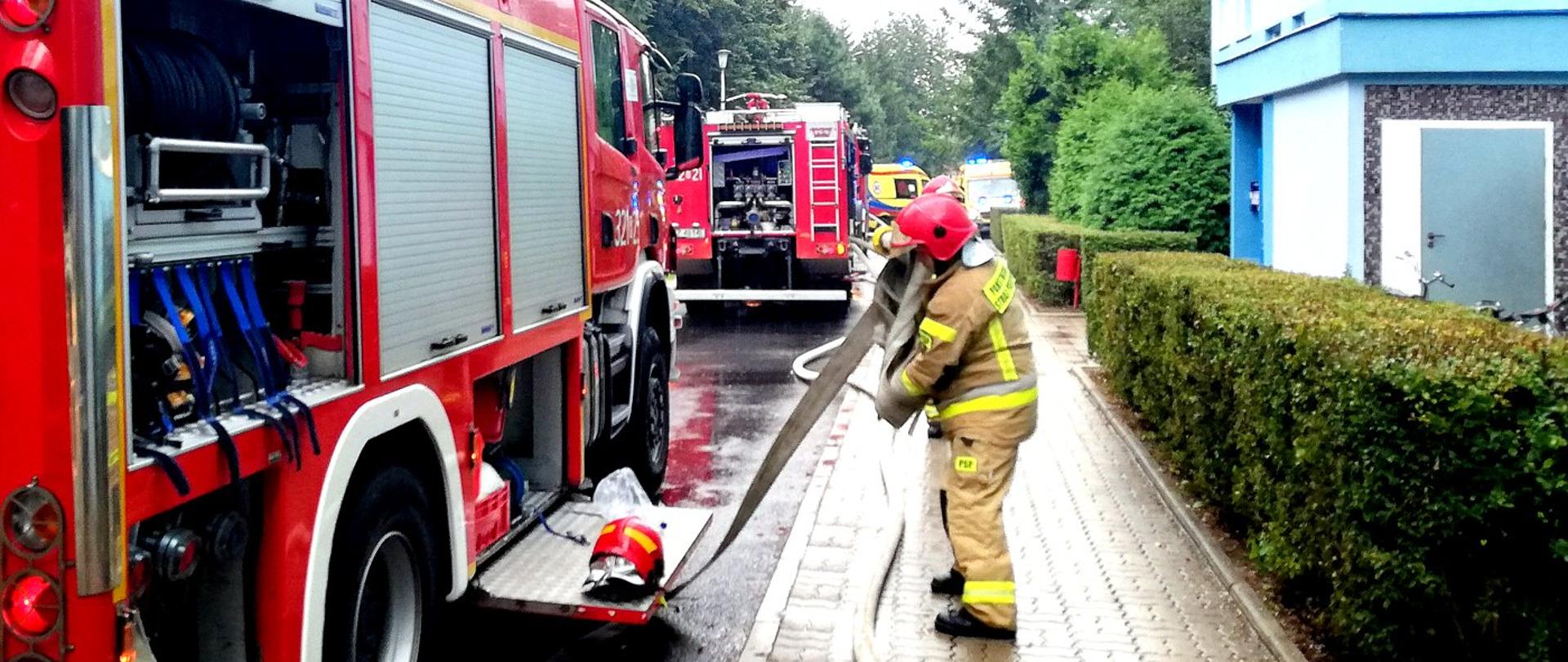 Na głównym planie strażak w mundurze bojowym zwija węża strażackiego. Po lewej stronie samochód strażacki z otwartymi skrytkami. W tle następny samochód strażacki oraz dwie karetki pogotowia. Po prawej stronie klatka schodowa budynku wielorodzinnego. 