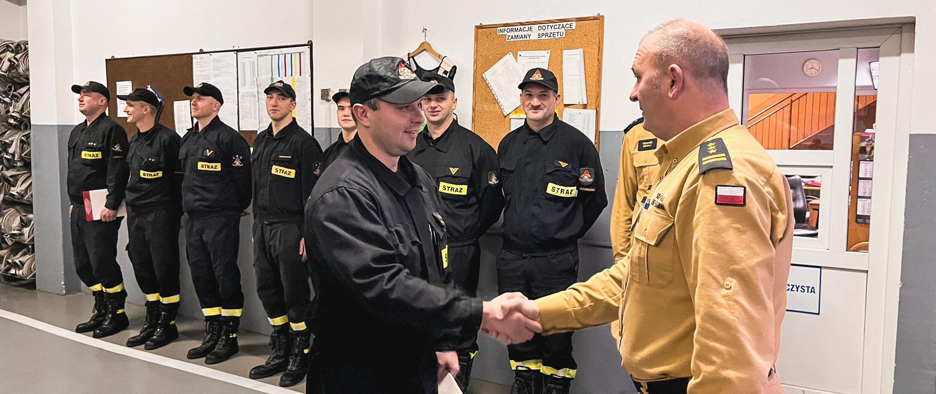 Strażak przyjmuje awans od komendanta ostrzeszowskich strażaków. Na drugim planie pozostali uczestnicy apelu. Zdjęcie wykonane wewnątrz strażnicy.