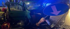 Rozbity pojazd osobowy na słupie oświetleniowym. Pora nocna. Po lewej stronie pojazdu widoczne sylwetki strażaków, za pojazdem zgromadzeniu ludzie i policjanci. W tle widoczny blok mieszkalny.