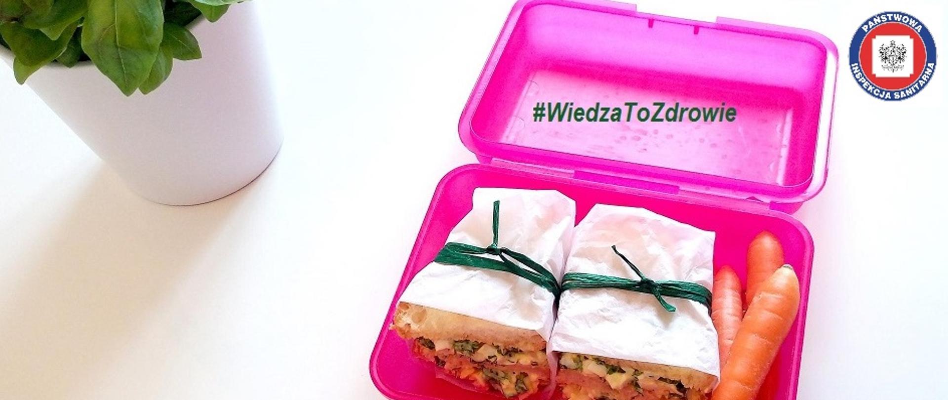 Na białym tle w lewym górnym rogu doniczka z zieloną bazylią, w prawym roku logo państwowej Inspekcji Sanitarnej. Po prawej stronie duży różowy otwarty kanapnik. W nim
dwie zawiązane "paczuszki" z kanapkami z warzywami, jajkiem, wędliną oraz trzy surowe marchewki. Na górnej części opakowania napis #WiedzaToZdrowie
