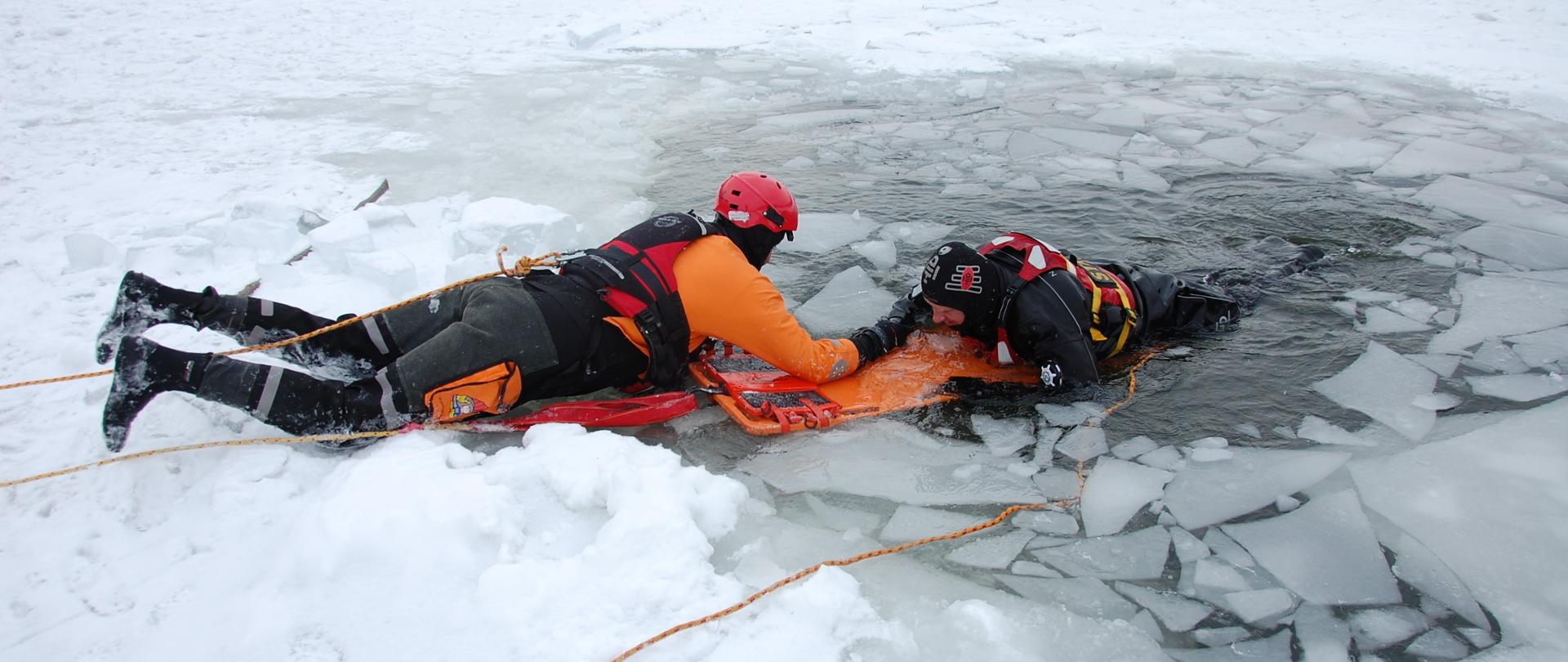 Na zdjęciu widzimy ratownika wydobywającego osobę poszkodowaną – pozoranta z przerębla z wykorzystaniem deski ortopedycznej. Wokół pozoranta widoczne są pokruszone kawałki tafli lodowej. Ratownik i pozorant są zaasekurowani pływającymi linami ratowniczymi.