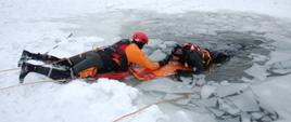 Na zdjęciu widzimy ratownika wydobywającego osobę poszkodowaną – pozoranta z przerębla z wykorzystaniem deski ortopedycznej. Wokół pozoranta widoczne są pokruszone kawałki tafli lodowej. Ratownik i pozorant są zaasekurowani pływającymi linami ratowniczymi.