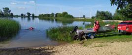 Samochód z łodzią stoi na jeziornej plaży a strażak podczepiony do linki płynie do pozoranta