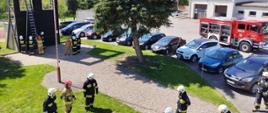 Zdjęcie przedstawia strażaków OSP podczas egzaminu n terenie przed Komendą. W tle wspinalnia i strażacy rozstawiający drabinę. po prawo wóz gaśniczy. w centrum zielona sosna.