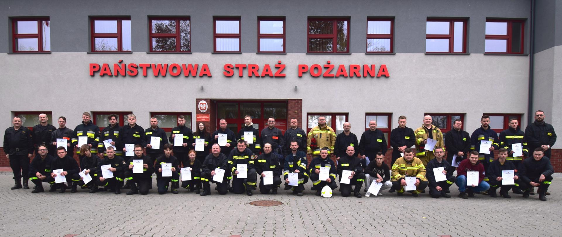 Grupa strażaków pozuje do zdjęcia po ukończonym szkoleniu. Strażacy ustawieni są w dwuszeregu, pierwszy szereg kuca, drugi stoi. W tle fragment budynku i napis Państwowa Straż Pożarna.