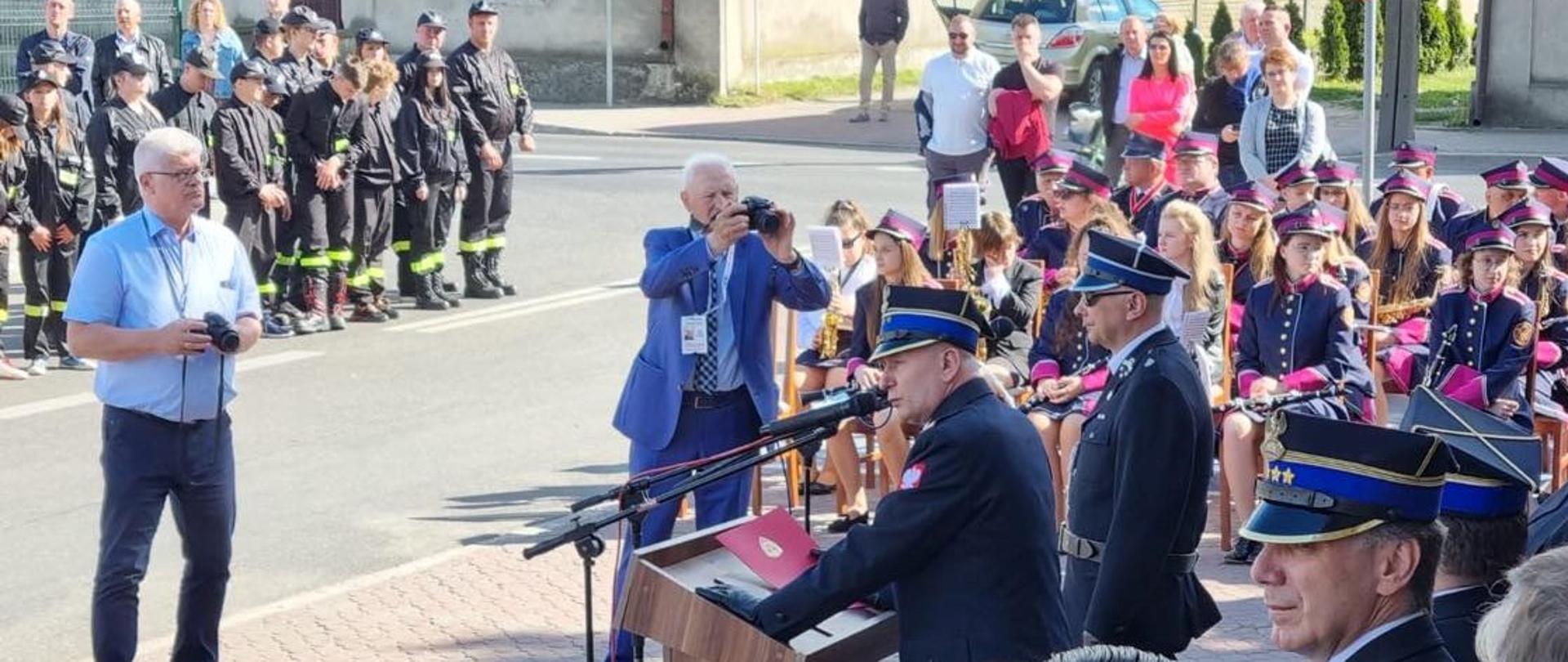 zastępca mazowieckiego komendanta wojewódzkiego psp przemawia podczas uroczystości. z boku widać orkiestrę dęta, po drugiej stronie stoją młodzi strażacy osp.